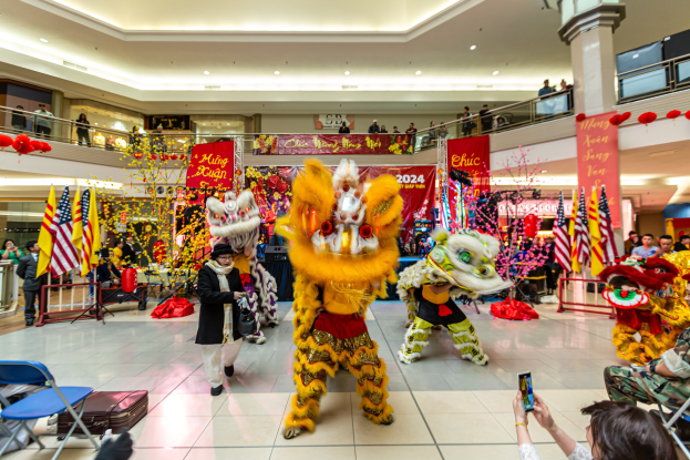Ein farbenfrohes chinesisches Neujahrsfest in einem Einkaufszentrum mit Menschen in Kostümen, die tanzen, anderen, die auf Stühlen sitzen und Handys benutzen, sowie Flaggen, Bannern, Dekorationsgegenständen, Geländern, Säulen, Deckenleuchten und einem Wasserzeichen unten.