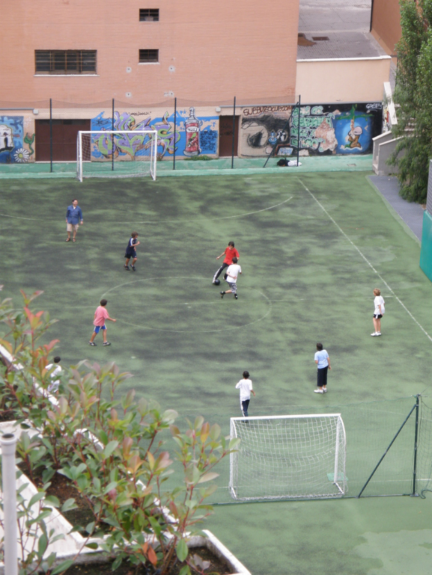 Kinder spielen Fußball auf einem Feld mit Toren, im Hintergrund ein Gebäude mit Graffiti, links Pflanzen und rechts Bäume.