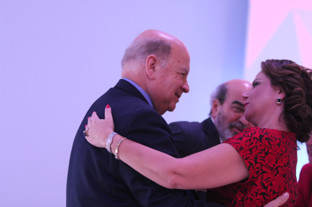 Zwei Menschen umarmen sich auf einer Bühne, umgeben von anderen, und einer trägt ein Armband.