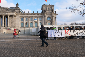 Eine Gruppe von Menschen marschiert vor dem Reichstaggebäude in Berlin, hält eine Fahne, mit dem Gebäude verziert mit Säulen, Fenstern, Bögen, Statuen und Flaggen, umgeben von einem Metallzaun, einem Straßenpfahl, einer Straßenlaterne, einigen Bäumen und einem bewölkten Himmel im Hintergrund.