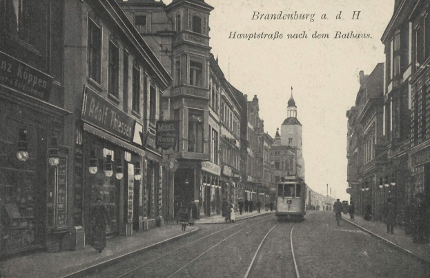 Ein altes Schwarz-Weiß-Foto einer Stadtstraße in Brandenburg, Deutschland, mit einer Straßenbahn auf den Schienen, Menschen auf dem Gehweg, Gebäuden mit Fenstern, Schildern, Straßenlaternen und einem bewölkten Himmel.
