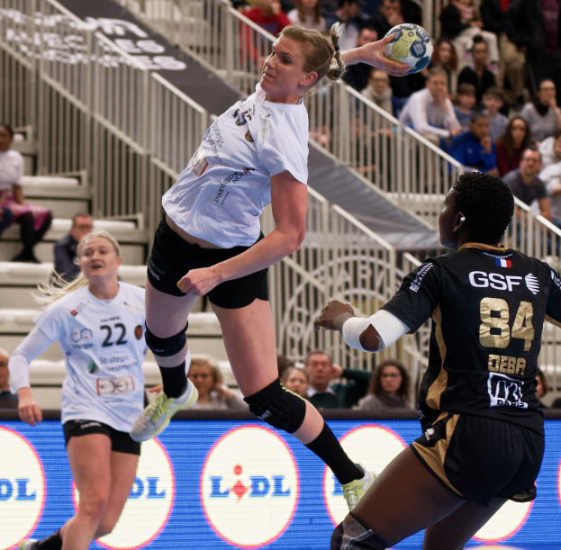 Gruppe von Frauen, die auf einem Platz Handball spielen, mit einem Ball in der Luft, Zuschauern auf Treppen und Geländern sowie einem "Handball-Weltmeisterschaft 2019 - Tag 2"-Schild im Hintergrund.