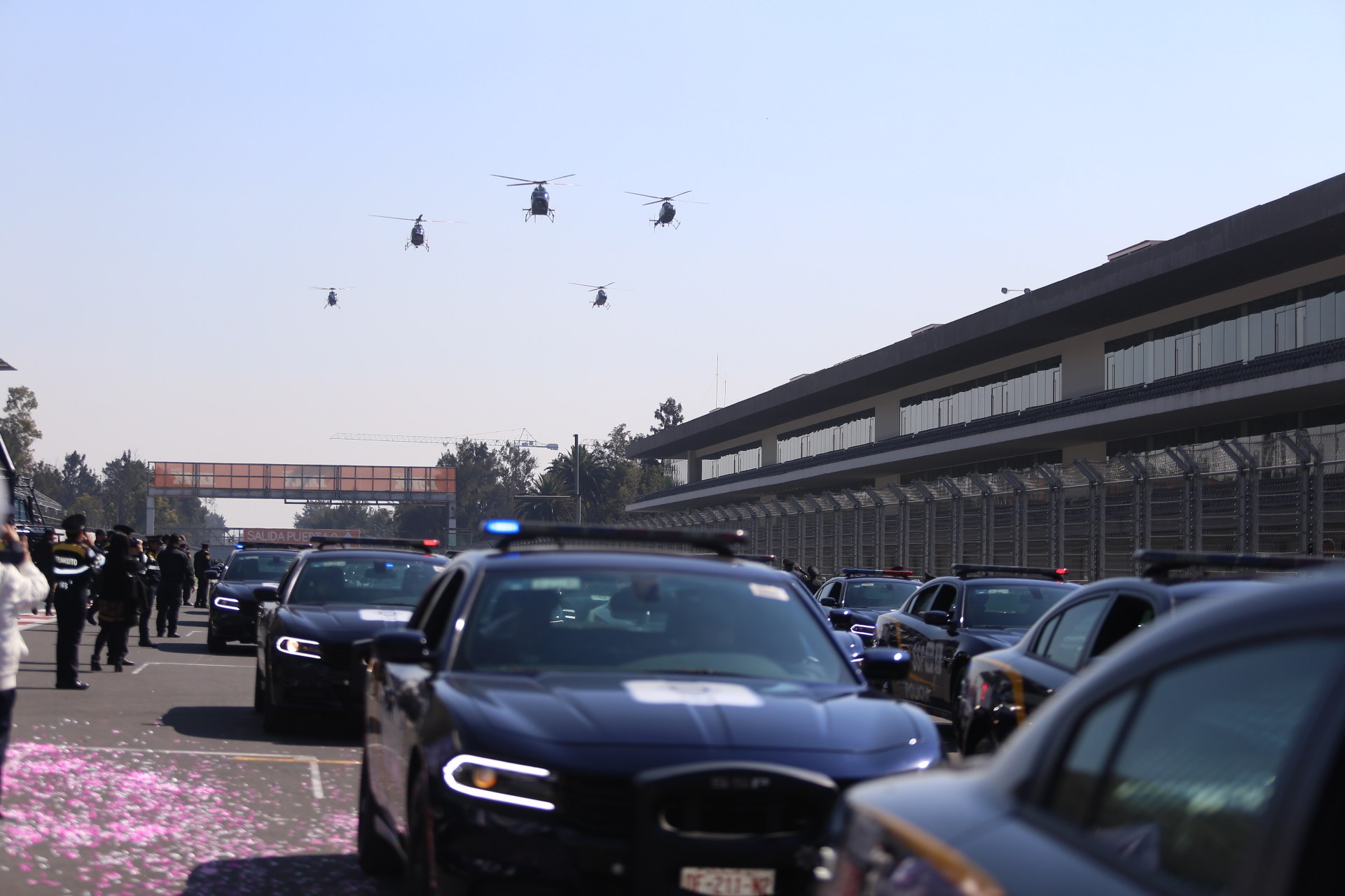 Polizeiautos vor einem Gebäude mit Menschen auf der linken Seite, Bäumen und einer Brücke im Hintergrund sowie vier Hubschraubern am Himmel.