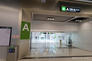 Ein leerer Flughafen-Terminal mit einer Wandtafel, die "Quarter Bld Nh" anzeigt, einem Mülleimer auf der linken Seite, einer Tafel mit Text und einigen Menschen im Hintergrund, sichtbarem Boden und Deckenbeleuchtung.