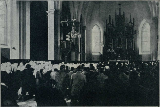 Ein Schwarz-Weiß-Foto einer großen Gruppe von Menschen, die in der Kirche des Heiligen Grabes in Jerusalem stehen, mit Säulen, Kronleuchtern und Fenstern im Hintergrund.