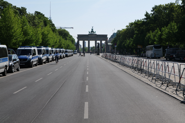 Eine Reihe von Polizeiwagen, die auf einer Straße vor dem Brandenburger Tor in Berlin, Deutschland, geparkt sind, mit Menschen auf Fahrrädern und auf der Straße stehend, Barrieren, Bäumen und einem Bogen mit Statuen im Hintergrund.