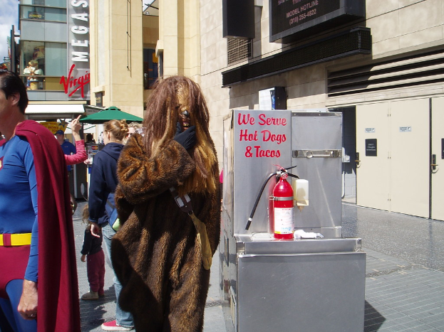 Eine Gruppe von Menschen, einige in Kostümen, versammelte sich vor einem Gebäude mit sichtbaren Türen und Beschriftung, mit einer Box und einem Feuerlöscher in der Nähe.