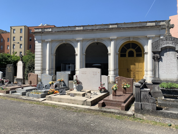 Friedhof mit zahlreichen Grabsteinen und Blumen, mit einem Gebäude mit gelber Tür, Bäumen, umliegenden Gebäuden mit Fenstern und einem klaren blauen Himmel.