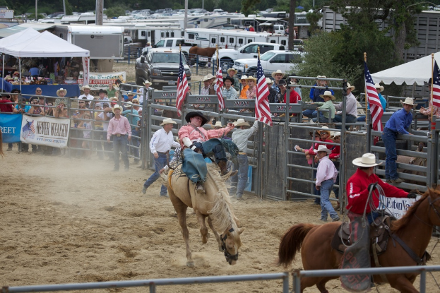 Zwei Reiter auf Pferden bei einem Wettbewerb namens Charreada, umgeben von Menschen, Fahnen und Zelten, parkenden Fahrzeugen und Bäumen im Hintergrund.