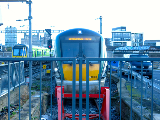 Zwei Züge auf den Schienen mit einem Zaun im Vordergrund, Pfähle mit einem mit einer Signalleuchte, ein Auto auf der Straße rechts, Gebäude im Hintergrund und Himmel oben.