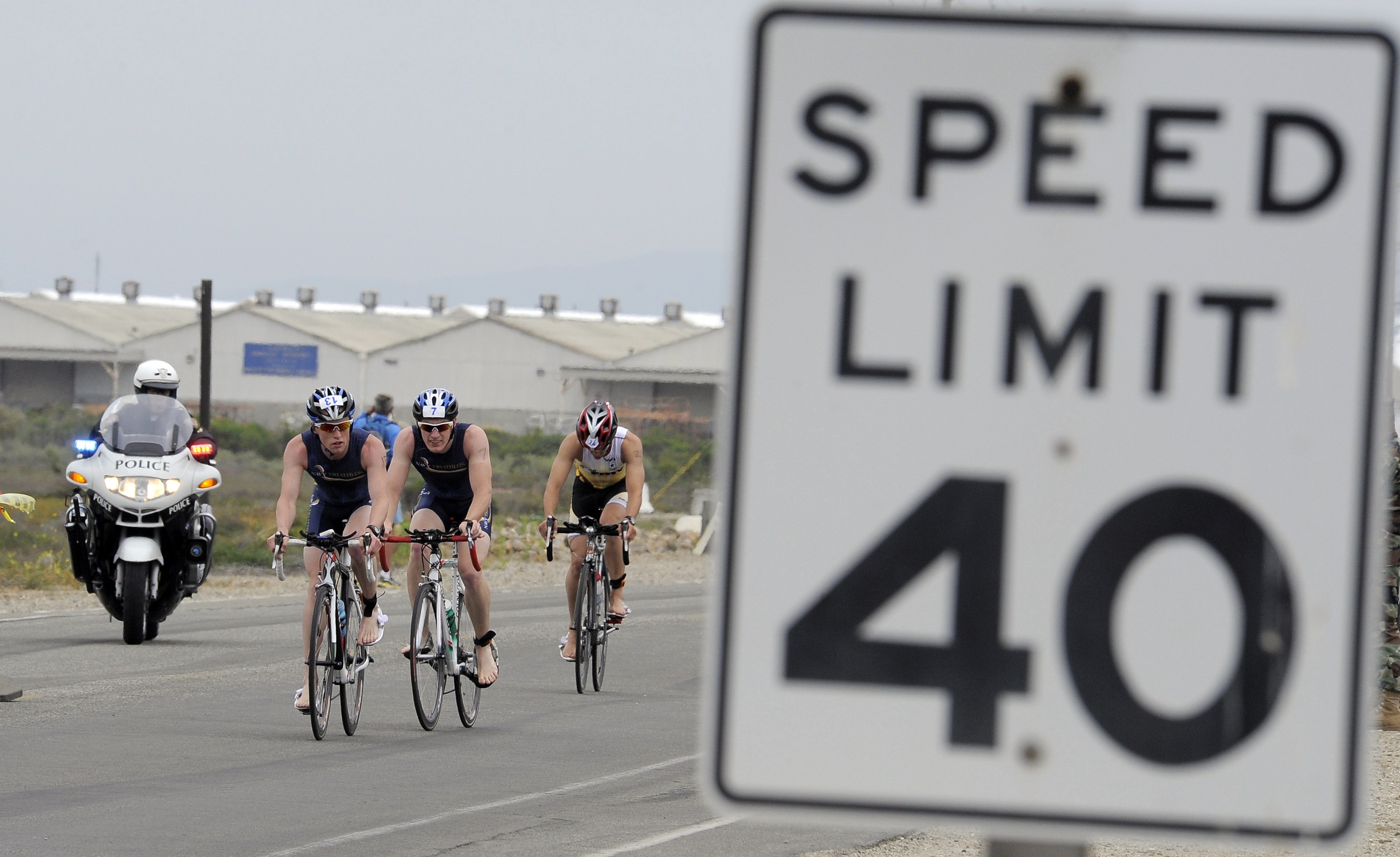 Gruppe von Menschen mit Helmen auf Fahrrädern auf einer Straße neben einem Geschwindigkeitsbegrenzungsschild mit Gebäuden, Pfählen, Pflanzen, Bäumen und einem klaren blauen Himmel im Hintergrund.