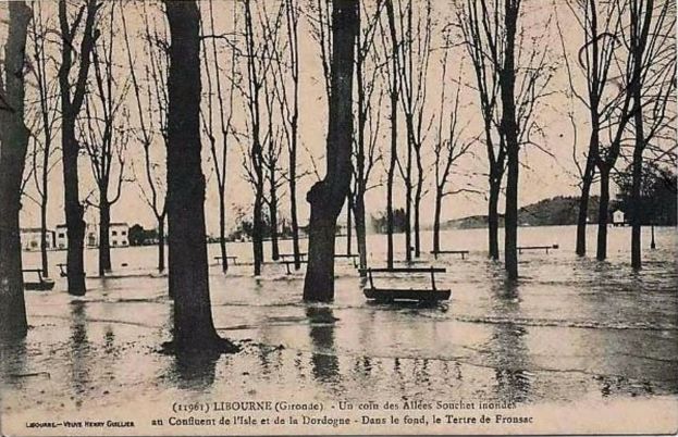 Ein Schwarz-Weiß-Foto eines überfluteten Parks mit Bäumen, Bänken, Wasser, Häusern, Hügeln und Himmel, mit Text unten.