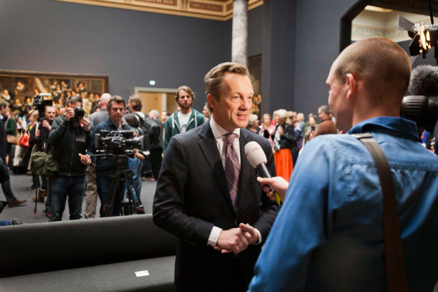 Ein Mann im Anzug und mit Krawatte spricht in ein Mikrofon, während er von einem Reporter interviewt wird, mit einer Menge Menschen im Hintergrund, einige halten Kameras, ein Sofa im Vordergrund und eine Wand mit einem Foto Rahmen, Lampen und einer Säule dahinter.