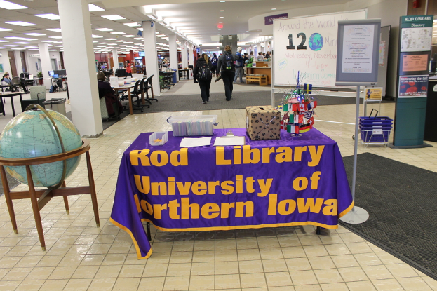 Eine Ausstellung mit einem Tisch, auf dem verschiedene Landesflaggen, eine Box, Papiere und ein Banner liegen, eine Kugel zur Linken, ein Anschlagbrett zur Rechten mit ein paar Menschen in der Nähe und mehreren Tischen und Stühlen im Hintergrund.