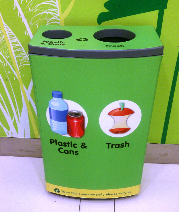 Ein grüner Recyclingmüllbehälter mit Plastik und Dosen auf einem weißen Boden mit einem Plakat an der Wand im Hintergrund.