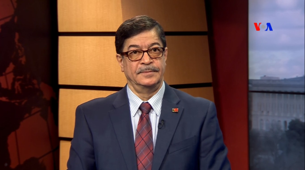 Ein Mann im Anzug und Krawatte sitzt vor einem Fernseher und trägt eine Brille mit ernster Miene.