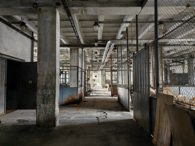 Leerer Raum mit großen Fenstern, Säulen, einer Tür, Deckenleuchten, Metallstangen, einem Maschendrahtzaun, verstreuten Gegenständen auf dem Boden und einer Wand, die eine verlassene Fabrik mit Anzeichen von Vernachlässigung zeigt.