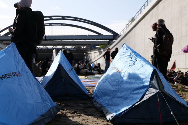 Eine Gruppe von Menschen sitzt auf einem sandigen Strand neben Zelten, mit einer Wand auf der rechten Seite und einer Brücke im Hintergrund, unter einem bewölkten Himmel, haltend Gegenstände und tragend Taschen während einer Klimawandel-Demonstration.