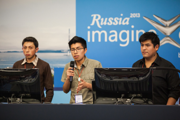 Drei Jungs stehen neben einem Schreibtisch mit zwei Computern, einer spricht in ein Mikrofon, alle tragen ID-Karten mit roten Markierungen, vor einer Wand mit einem Werbeplakat, auf dem "Russia Imagine 2013" steht.