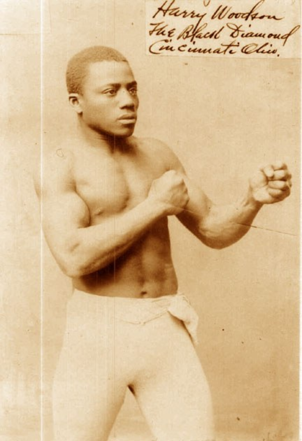 Ein Schwarz-Weiß-Foto eines Mannes in einem Boxring mit Handschuhen und Shorts, der mit konzentrierter Miene steht.