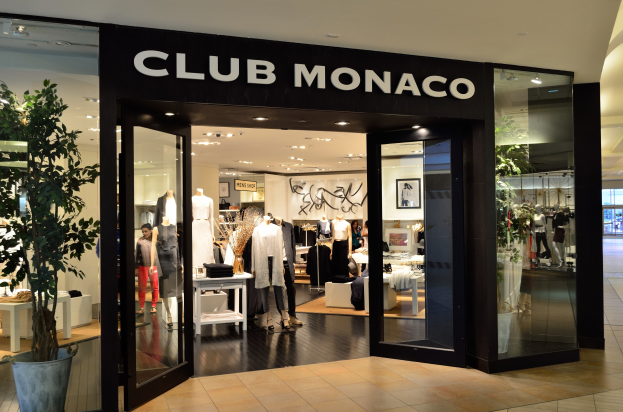 Außenansicht eines Club Monaco-Geschäfts in einem Einkaufszentrum mit Glaswänden, Schaufensterpuppen in Kleidern, Ausstellungstischen, einem Topf mit einer Pflanze und Deckenbeleuchtung.