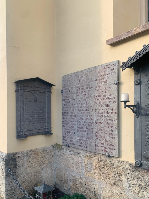 Ein Geb├Ąude mit einer Gedenktafel f├╝r Holocaust-Opfer, mit einem Fenster, einer T├╝r, Licht, Kette und nahen Pflanzen.