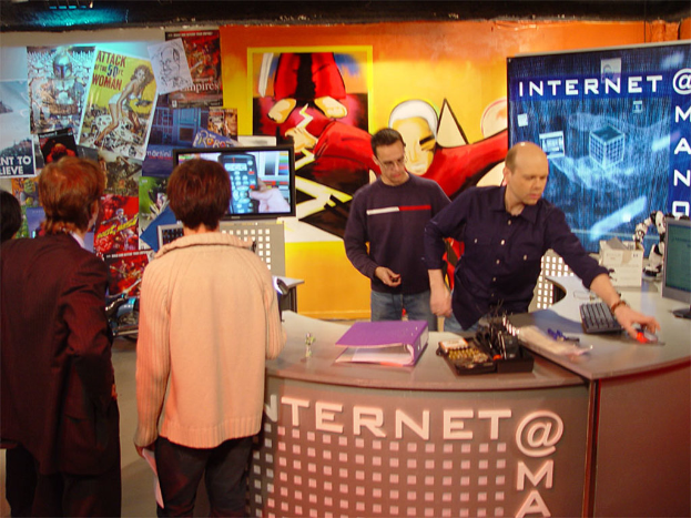 Mehrere Personen in einem Raum mit einem Tisch, auf dem sich eine Akte, eine Tastatur, ein Monitor und eine Maus befinden, sowie eine Wand im Hintergrund mit Plakaten und einer Cartoon-Figur auf dem Bildschirm.