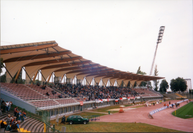 Altes Schwarz-Weiß-Foto eines Stadions voller Zuschauer, das sitzende Fans unter der Dachkonstruktion, ein geparktes Auto auf dem Feld, stehende Besucher auf dem Rasen, Stangen und Rahmen, umliegende Zäune, eine Straßenlaterne, entfernte Bäume, einen hohen Turm und einen bewölkten Himmel zeigt.