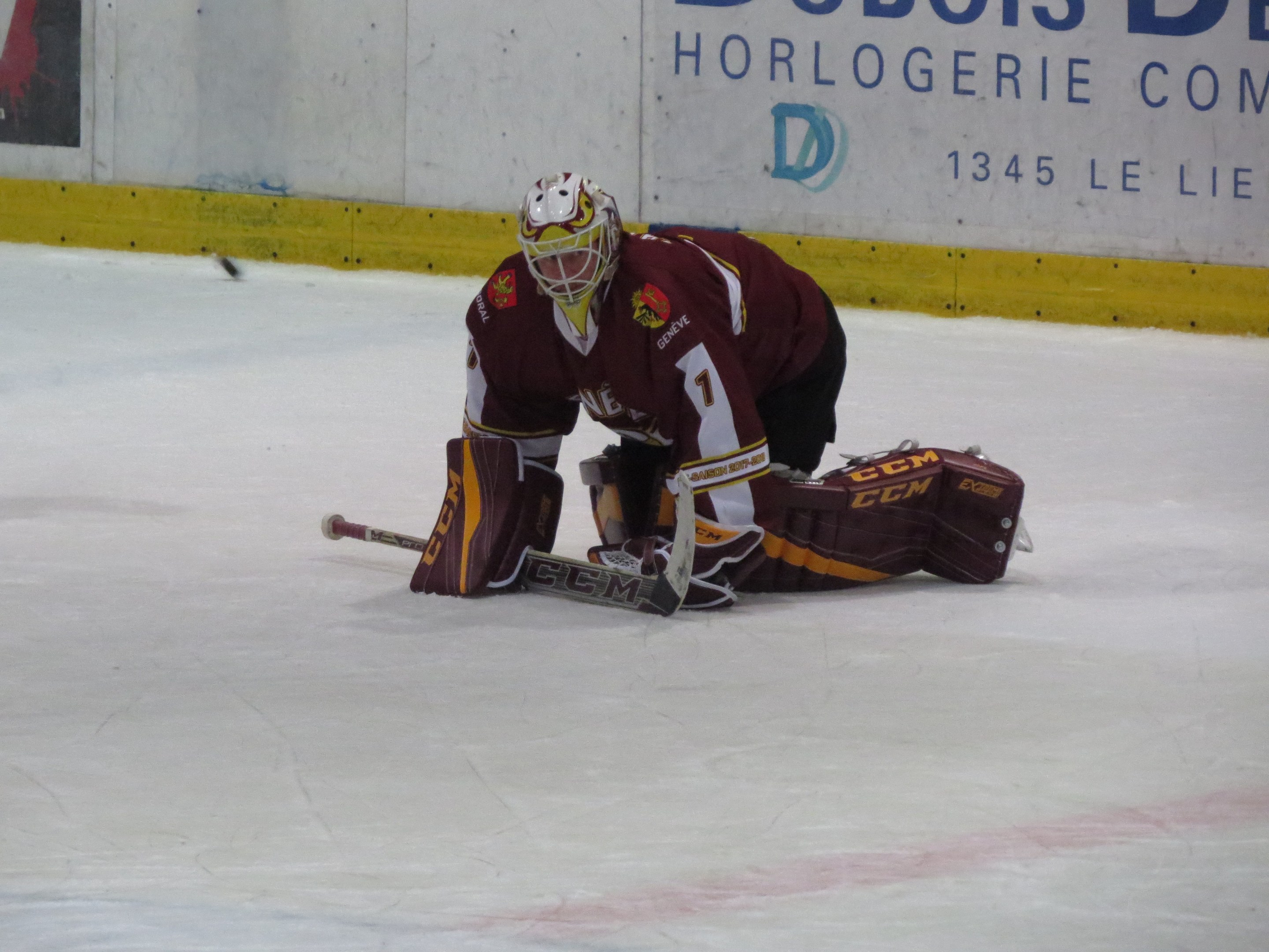Eishockeyspieler in rot-gelber Uniform, der einen Schuss hält, trägt Helm, Handschuhe und Knieprotektoren und hält einen Hockey-Schläger, mit einer Wand und Text im Hintergrund.