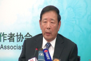 Ein Mann im Anzug sitzt an einem Tisch mit Mikrofonen und spricht während einer Presseveranstaltung mit einem Banner im Hintergrund.