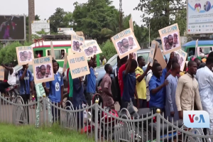 Eine Gruppe von Menschen hält Schilder und steht vor einem Zaun, umgeben von Fahrzeugen, Bäumen, Pfosten, Werbetafeln und einem Bildschirm, mit Gras am Boden und dem Himmel im Hintergrund, wahrscheinlich protestieren sie gegen eine Regierungsentscheidung, die Video-Konferenzen verbietet.