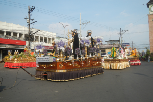 Ein Paradewagen mit Menschen darauf, umgeben von Gebäuden, Strommasten mit Drähten, Laternenmasten, Bäumen und einem Kirchturm im Hintergrund unter einem bewölkten Himmel.