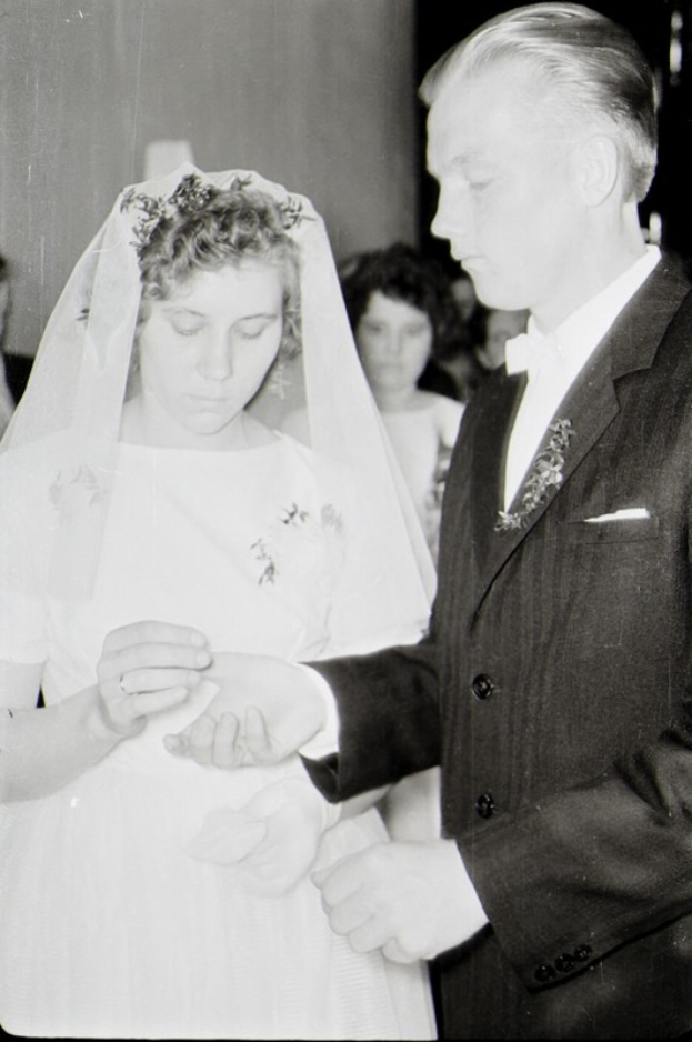 Ein Schwarz-Weiß-Foto einer Braut in einem weißen Kleid und Schleier und eines Bräutigams in einem eleganten Anzug beim Austausch der Eheringe, mit einigen Leuten im Hintergrund.