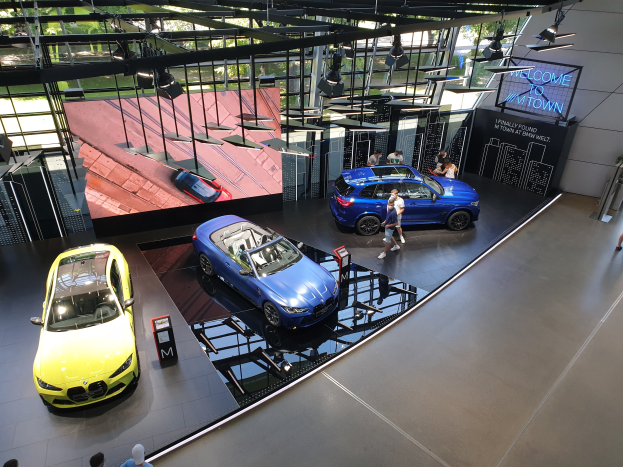 Eine Gruppe von Menschen im BMW Museum in München, Deutschland, mit Autos zur Linken und einem Schild mit der Aufschrift "BMW Museum München" zur Rechten, mit Blick auf eine Außenansicht von Bäumen, Gebäuden und Himmel durch Glasfenster.