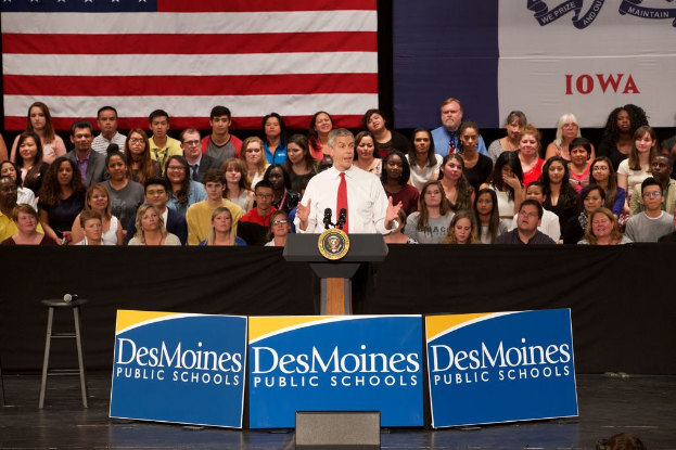 Ein Mann in weißem Hemd und roter Krawatte steht an einem Podium mit drei Texttafeln, einem Hocker mit Mikrofon und einer sitzenden Menge im Hintergrund, mit einer Flagge und zusätzlichem Text.