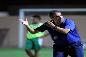 Ein Mann in einem blauen T-Shirt und Hose steht auf einem Fußballfeld und hält ein Mikrofon in der Hand, während er auf seine Spieler zeigt. Im Hintergrund sind eine Person in einem grünen Kleid, ein Torpfosten und eine Wand zu sehen.