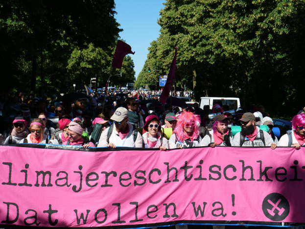 Eine Gruppe von Menschen marschiert mit einem pinken Banner mit der Aufschrift 'Frauenmarsch in Berlin' die Straße entlang, einige tragen Mötze und Schutzbrillen, andere halten Fahnen, vor dem Hintergrund von Bäumen, Fahrzeugen und Himmel.