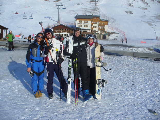Vier Personen posieren mit Skaterausrütstücken und Werkzeugen für ein Foto, mit Gebäuden und anderen Menschen im Hintergrund, vor einer schneebedeckten Berglandschaft.