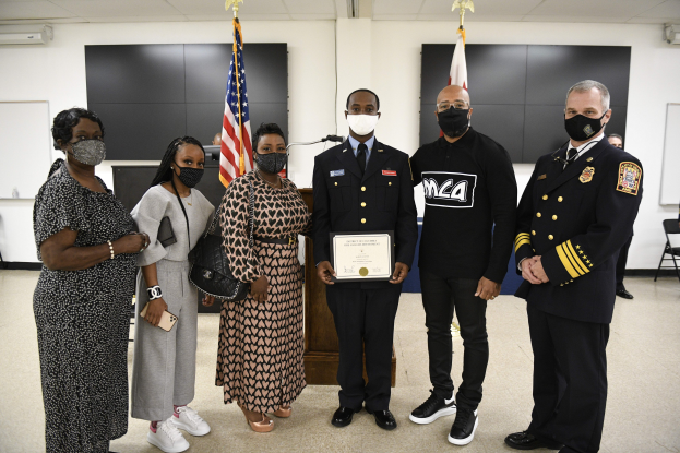 Eine Gruppe von Menschen mit Masken steht zusammen vor Flaggen, wobei die zentrale Person ein Zertifikat hält, während einer Abschlussfeier in einem Raum mit Stühlen, Tischen und Wandtafeln.