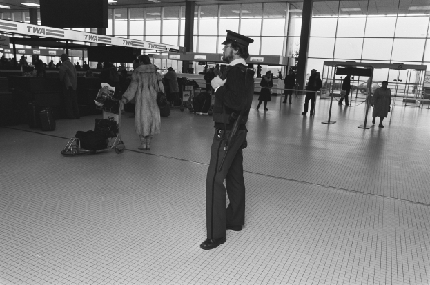 Ein Polizist in Uniform steht in einem Flughafen-Terminal und h├Ąlt eine Waffe in der Hand, umgeben von Menschen und Gep├Ąckwagen, mit Masten, Texttafeln, Deckenleuchten und Glasw├Ąnden im Hintergrund.