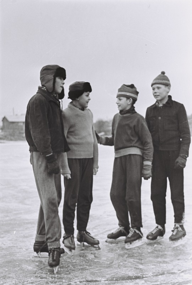 Vier junge Jungs in Mützen und Schlittschuhschuhen stehen auf einem zugefrorenen See, mit Häusern, Bäumen und einem klaren Himmel im Hintergrund, dargestellt in Schwarz-Weiß.