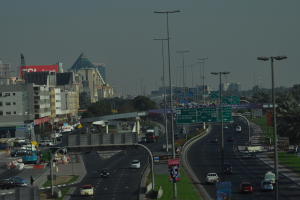 Eine Luftaufnahme einer Stadt mit Gebäuden, Türmen, Kraftfahrzeugen, Werbetafeln, Schildern, Straßen, Bäumen, Straßenlaternen, Menschen, Verkehrsleitkegeln und Informationsanzeigen unter einem klaren Himmel.