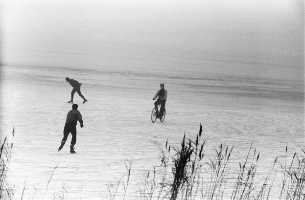 Drei Menschen auf einem zugefrorenen See Schlittschuhlaufen, einer mit Fahrrad, umgeben von Gras, in Schwarz-Weiß.