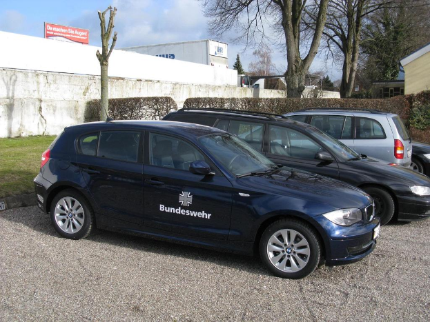 Mehrere Autos stehen auf einem von grünem Gras, Büschen und Bäumen umgebenen Weg. Im Hintergrund sind Gebäude, ein Schild mit Text und ein sichtbares Logo zu sehen.