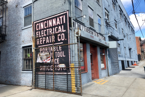 Das Gebäude der Cincinnati Electrical Repair Co. mit Fenstern, Türen, einem Schild, einem Metallzaun, einem Müllcontainer, Polen, Drähten, Bäumen und einem bewölkten Himmel.