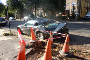 Ein verunglücktes Auto am Straßenrand, umgeben von orangen Verkehrskegeln, mit Bäumen, Gebäuden und einem klaren blauen Himmel im Hintergrund.