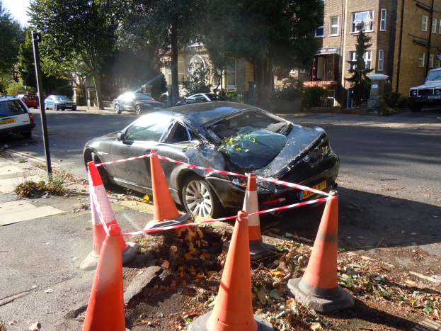 Ein verunglücktes Auto am Straßenrand, umgeben von orangen Verkehrskegeln, mit Bäumen, Gebäuden und einem klaren blauen Himmel im Hintergrund.