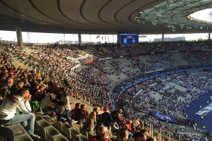 Große Menschenmenge in einem Stadion bei einem Fussballspiel mit einer Bühne, Fahnen, Stangen und einem Bildschirm im Hintergrund, wahrscheinlich die Allianz Arena in München, Deutschland.