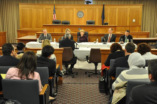 Ein Versammlungsraum mit einer Gruppe sitzender Menschen, zwei ausländischen Flaggen, einem orangefarbenen Schreibtisch mit Wasserflaschen und einem Mikrofon.