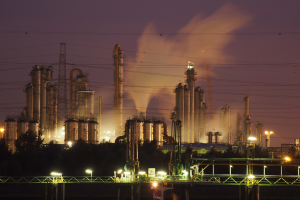 Ölraffinerie bei Nacht mit Rauch, beleuchtet von umliegenden Lichtern mit sichtbarem Himmelhintergrund und Wasserzeichen.